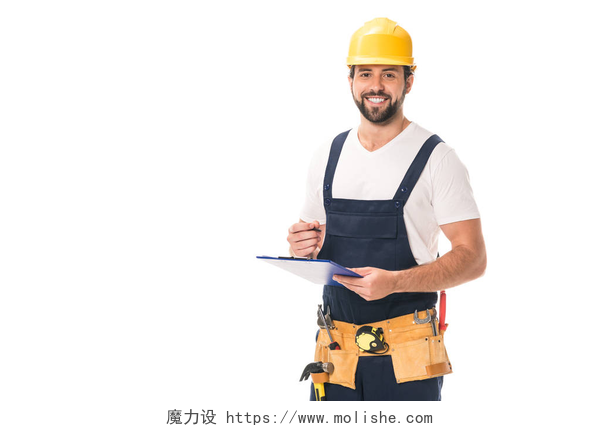 在白色背景墙前一个在记录的工人英俊快乐的工匠在工具带和硬帽子写在剪贴板上, 微笑着在白色的相机隔离